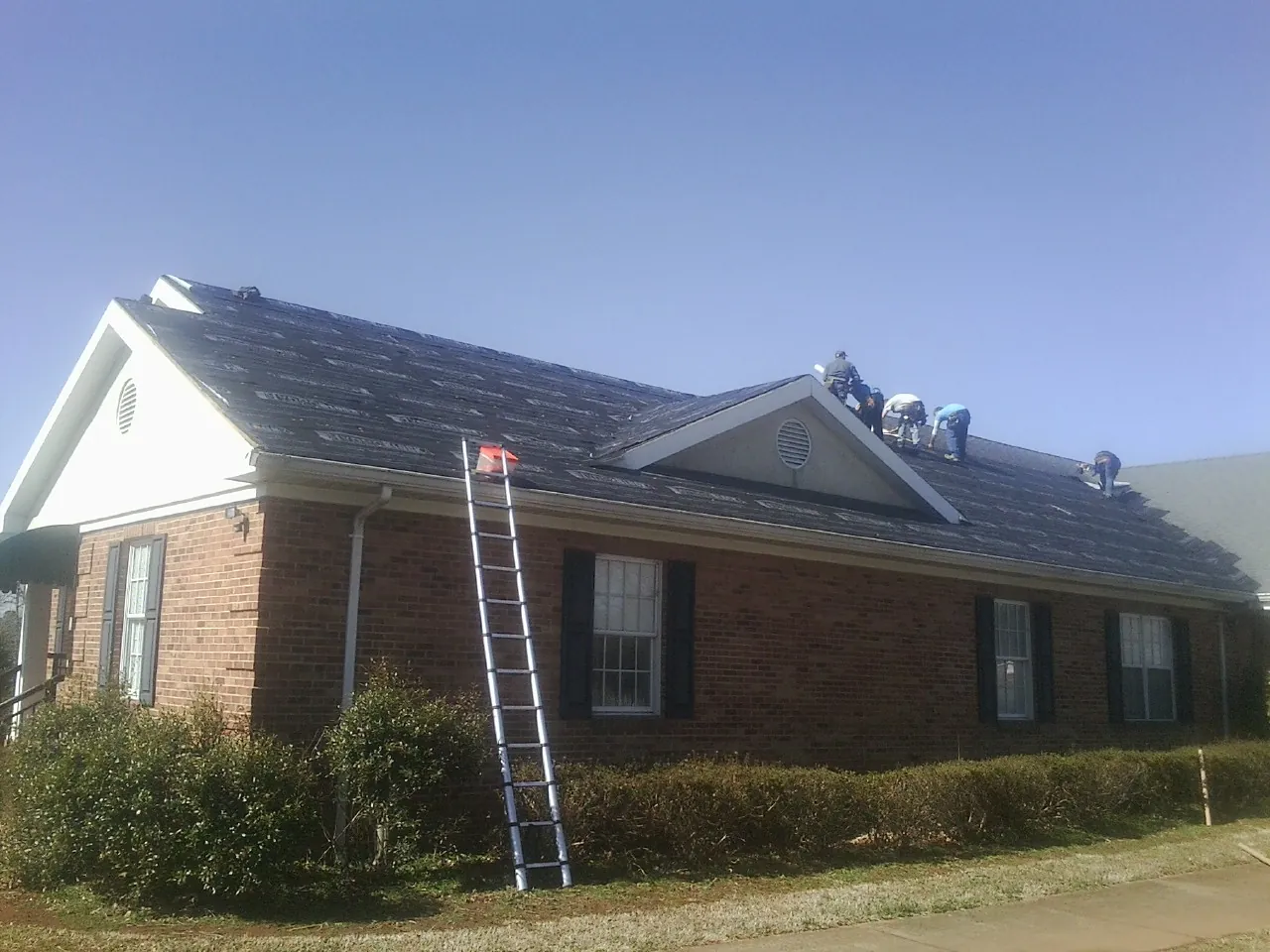 Crew tearing shingles off a brick ranch roof with a ladder set — Emergency Roof Repair work by our crew in Saluda