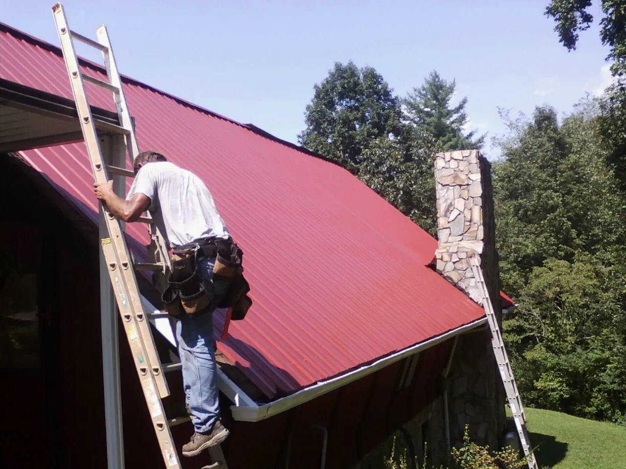 Roofer on a ladder beside a red metal roof and stone chimney — Soffit & Fascia Repair by Roofing Brothers in Saluda